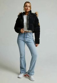 Jachetă matlasată neagră, scurtă, cu blană la margine, cu fermoar cu model, asortată cu blugi evazați bleu deschis și adidași albi.