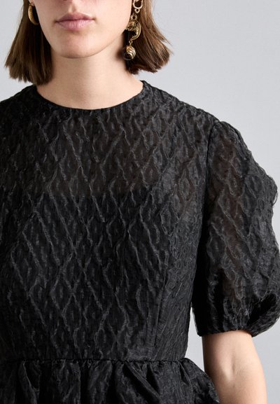 Woman wearing textured black puff sleeve dress and gold sculptural earring, shown from chin to waist against a plain gray background.