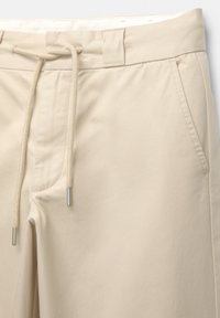 Chinos beige avec une taille à cordon, comportant deux poches latérales et une texture lisse. Le tissu est doux et léger.