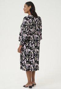 Vestido midi negro con estampado abstracto en rosa y crema, cintura elástica, mangas largas y un corte relajado; combinado con sandalias negras de tacón cuadrado.