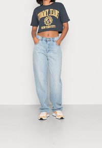 Šedé zkrácené tričko s potiskem "TOMMY JEANS NEW YORK CITY", zkombinované se světle modrými vysokými džíny s rovnýma nohavicema a bílými-oranžovými teniskami.