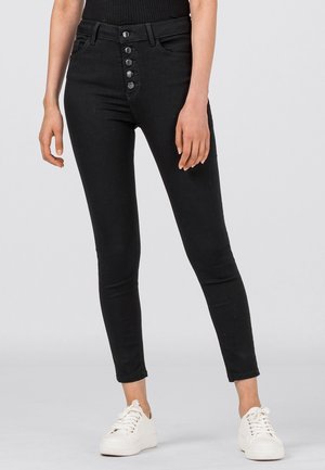 Femme portant un jean skinny noir taille haute avec fermeture à cinq boutons et des baskets blanches, debout sur un fond uni.