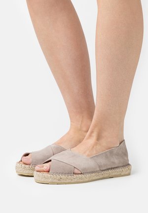 Personne portant des sandales espadrilles en suède beige avec des brides croisées et des semelles en jute tressé sur fond blanc.