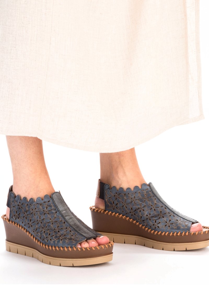 Sandales compensées avec une tige en cuir bleu marine, ornée de découpes florales, d'un bord marron et d'une semelle profilée. Le design ouvert à l'avant met en valeur les orteils.