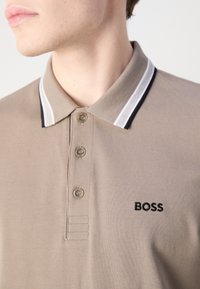 Gros plan d'une personne portant un polo beige avec un col rayé et le logo « BOSS » sur la poitrine.