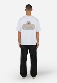 Mężczyzna w białej koszulce z kolorową grafiką "Pegador Whispers of Cold" na plecach, w luźnych czarnych spodniach i jasnych butach, stojący tyłem.