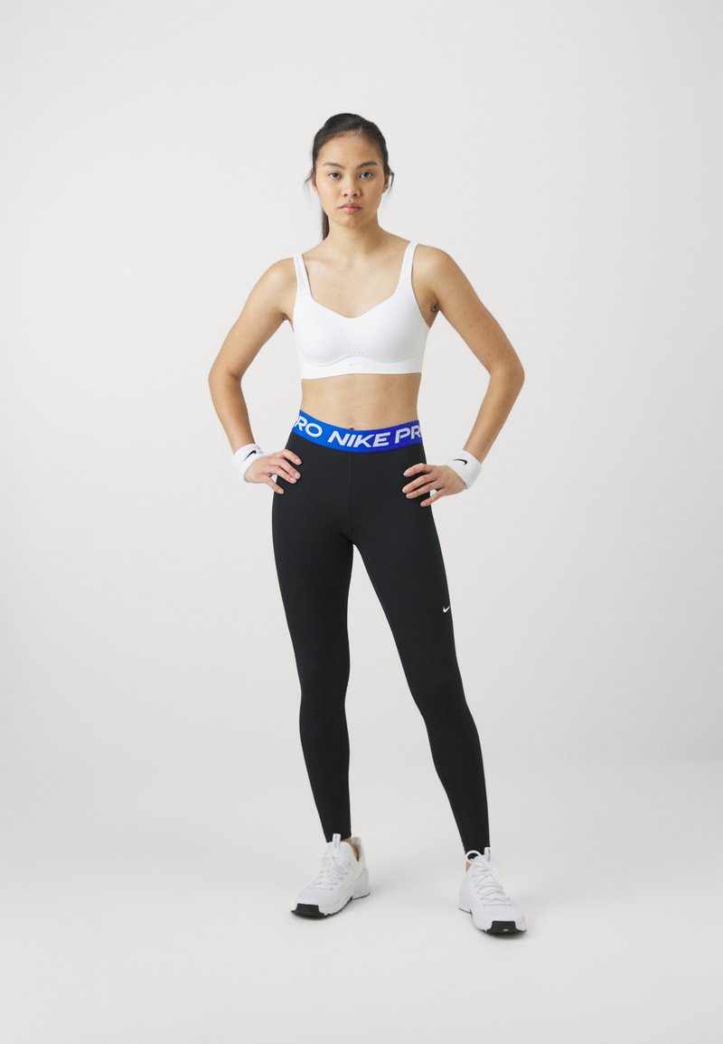 Leggings noires avec ceinture "PRO NIKE" bleue, soutien-gorge de sport blanc, et baskets blanches ; le modèle porte des bracelets et a les cheveux raides.