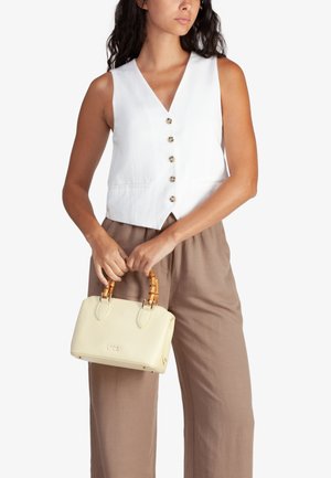 Gilet bianco senza maniche con bottoni, abbinato a pantaloni a gamba larga beige chiaro. Tiene in mano una piccola borsa color crema con finitura testurizzata e manici in bambù.