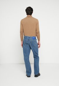 Καφέ μακρυμάνικη μ�πλούζα με εφαρμοστή σχεδίαση· μπλε τζιν παντελόνι με ίσια γραμμή, εμφανείς ραφές και ετικέτα με λογότυπο στη μέση.