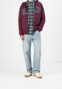 Sudadera con capucha granate con el logo "GAP", superpuesta sobre una camisa de cuadros verde. Vaqueros anchos azul claro y zapatillas crema con suelas marrones claras.