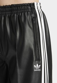 Svarta faux läderbyxor med vita ränder på sidorna och en liten vit Adidas-logga på framsidan. Har en elastisk midja och ficka.
