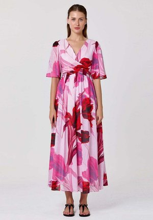 Femme portant une robe portefeuille fleurie rose et rouge à manches mi-longues et des sandales noires sur un fond clair uni.