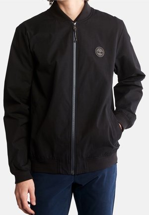 Person trägt eine schwarze Jacke mit Reißverschluss und einem kleinen Baum-Logo auf der Brust sowie marineblaue Hose, eine Hand in der Tasche.