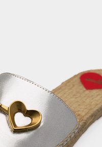 Ασημένια σανδάλι με φαρδιά σόλα από πλεγμένο υλικό και μεταλλικό διακοσμητικό με σχήμα καρδιάς σε χρυσό χρώμα στο λουράκι, με κόκκινη ετικέτα στη σόλα.