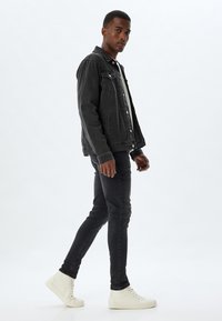 Chaqueta de denim negra con cierre de botones y dos bolsillos en el pecho, combinada con jeans skinny negros y zapatillas altas blancas.