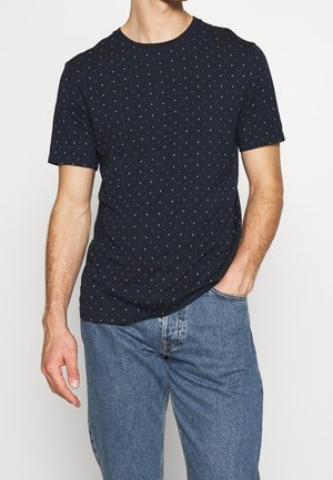 Hombre que lleva una camiseta de manga corta azul marino con pequeños lunares blancos y unos vaqueros de denim azul claro, con una mano en el bolsillo.