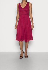 Robe mauve ajustée à la taille avec un corsage en dentelle, un décolleté en V et une jupe fluide ; associée à des escarpins noirs à talons aiguilles et à des sandales lisses couleur peau.