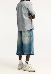 Camisa gris claro de manga corta combinada con pantalones cortos de mezclilla azul descoloridos hasta la rodilla, que cuentan con dos bolsillos traseros y un dobladillo deshilachado. Zapatillas blancas y calcetines.