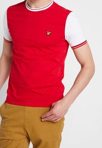 Camiseta roja de manga corta con mangas blancas, cuello acanalado a rayas y pequeño logo verde. Material de algodón con una textura suave.