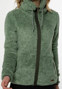 Polarowa kurtka w stonowanym zielonym kolorze, z wysokim kołnierzem, pełnym zamkiem błyskawicznym, sznurkami oraz fakturowanym materiałem, z bocznymi kieszeniami i kontrastowymi detalami.