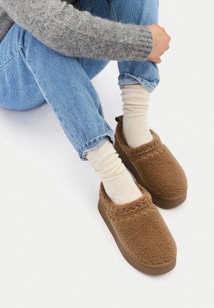 Persona seduta con le gambe incrociate che indossa jeans blu, calzini color crema e pantofole marroni pelose senza lacci con suola spessa su sfondo bianco.