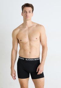 Oberkörperfreier Mann mit kurzen braunen Haaren, der schwarze Björn Borg Boxershorts trägt und vor einem einfarbigen, hellen Hintergrund steht.