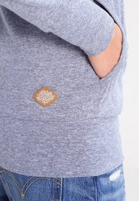Grauer, strukturierter Sweatshirt mit einer Seitentasche und einem braunen, rautenförmigen Etikett mit weißem Blumendesign, kombiniert mit Jeans.