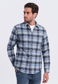 Camisa de franela para hombre en cuadros azules y grises. Cuenta con cuello abotonado, mangas largas y un bolsillo en el pecho. Tejido suave y texturizado.