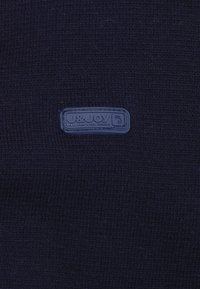 Marineblauwe gebreide stof met een gestructureerd oppervlak. Inclusief een blauw rechthoekig logo-patch met "J&Joy" en "Elke stap een nieuwe reis" in reliëf.