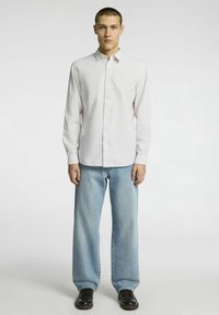 Balta marškiniai su ilgomis rankovėmis, derinami su šviesiai mėlynomis tiesiomis kelnėmis ir juodais užrišamais batais. Minimalus tekstūros matomumas.