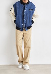 Kurtka typu bomber z denimu z kremowymi rękawami, czarnymi ściągaczami i wyhaftowanym logo „HUGO”; w zestawie beżowe spodnie i białe trampki.