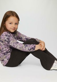 Lila Langarmshirt mit floralem Muster, kombiniert mit schwarzen Hosen. Das Kind sitzt mit übergeschlagenen Beinen und trägt weiße Turnschuhe.