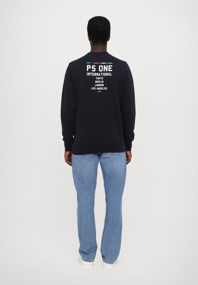 Mustat collegepaita, jossa on graafinen teksti selässä, yhdistettynä vaaleansinisiin farkkuihin. Resorikankaiset hihansuut ja helma. Materiaali vaikuttaa pehmeältä ja rentolta.