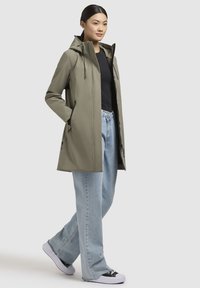 Giacca impermeabile verde oliva chiaro con cappuccio, dotata di tasche laterali, cordini regolabili e un design liscio e elegante. Abbinata a jeans a gamba larga e sneaker.