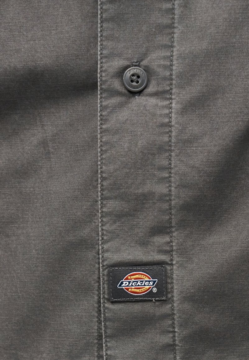 Tessuto grigio con cuciture verticali, bottone nero centrale con rilievo "Dickies" e una piccola toppa rettangolare nera con il logo Dickies sotto.