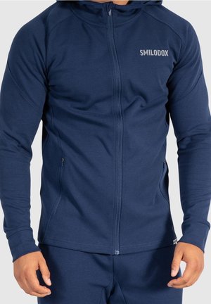 Man draagt een aansluitende marineblauwe hoodie met rits en het logo "SMILODOX" op de borst, gecombineerd met bijpassende marineblauwe broek, tegen een effen achtergrond.