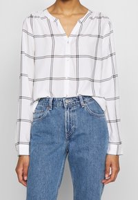 Femme portant une chemise blanche à manches longues avec un motif à carreaux noirs, rentrée dans un jean bleu taille haute, les mains détendues sur les côtés.