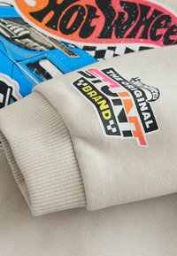 Primer plano de la manga de una sudadera beige con el logo de Hot Wheels Stunt Brand y un gráfico colorido de Hot Wheels en la tela.