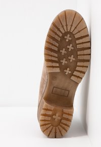 Semelle d'une botte beige montrant des crampons profonds avec des rainures horizontales et des motifs en forme de croix, sur un fond blanc, sol et mur.