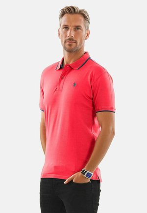 Rotes Poloshirt mit marineblauen Kragen und Ärmelakzenten, aus Baumwolle, klassischer Schnitt, mit gesticktem Logo auf der Brust, kombiniert mit schwarzen Hosen.