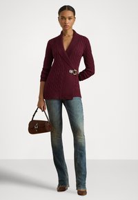 Burgunderfarget strikket cardigan med omslagsdesign og spenne-detalj, kombinert med falmede denimjeans og en liten brun semsket skinnveske.