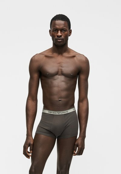 Μυώδης άνδρας που στέκεται στραμμένος προς τα εμπρός, φορώντας γκρι μπόξερ Emporio Armani, με απλό φωτεινό φόντο.