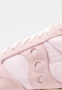 Gros plan sur une sneaker en daim rose clair et en tissu, avec des lacets blancs tressés et un design circulaire brodé sur le côté.