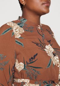 Nærbillede af en person, der bærer en brun blomstrete skjorte med grønne blade og cremede blomster samt små guld hoop-øreringe.