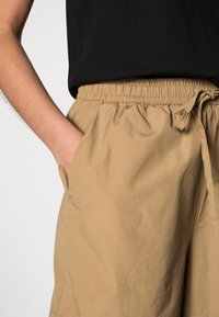 Beige shorts, valmistettu kevyestä kankaasta, jossa on joustava vyötärö ja nyöri, sivutaskut ja sileä pinta.