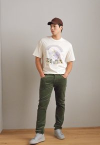 Bela grafična majica s napisom "LEE JEANS", olivno zeleni hlače in bele športne copate. Rjava bejzbol kapa. Stoječi položaj z nevtralnim izrazom.