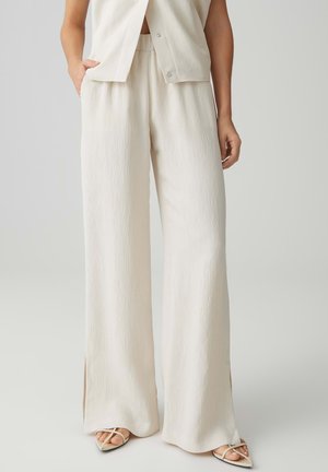 Vrouw draagt losse, crèmekleurige, gestructureerde wijde broek met zijsplitten, gecombineerd met een bijpassende mouwloze top en witte sandalen met bandjes en hak.