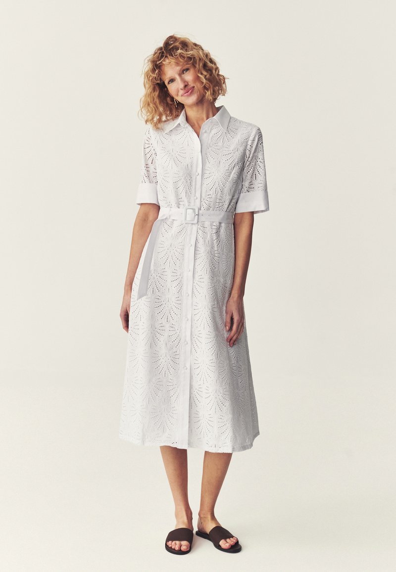 Femme aux cheveux bouclés portant une robe-chemise midi blanche à œillets, manches courtes, une ceinture et des mules noires à bout ouvert, debout sur un fond uni.