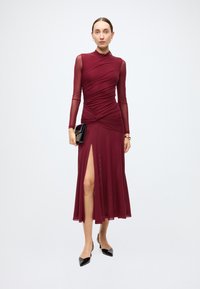Burgundy dress with long sheer sleeves, ruched bodice, and a high slit. Styled with black accessories and pointed-toe heels. Smooth, flowing fabric.