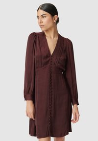 Femme aux cheveux bruns en chignon bas portant une robe à manches longues de couleur bordeaux profond avec des boutons sur le devant et des plis au niveau de la poitrine.